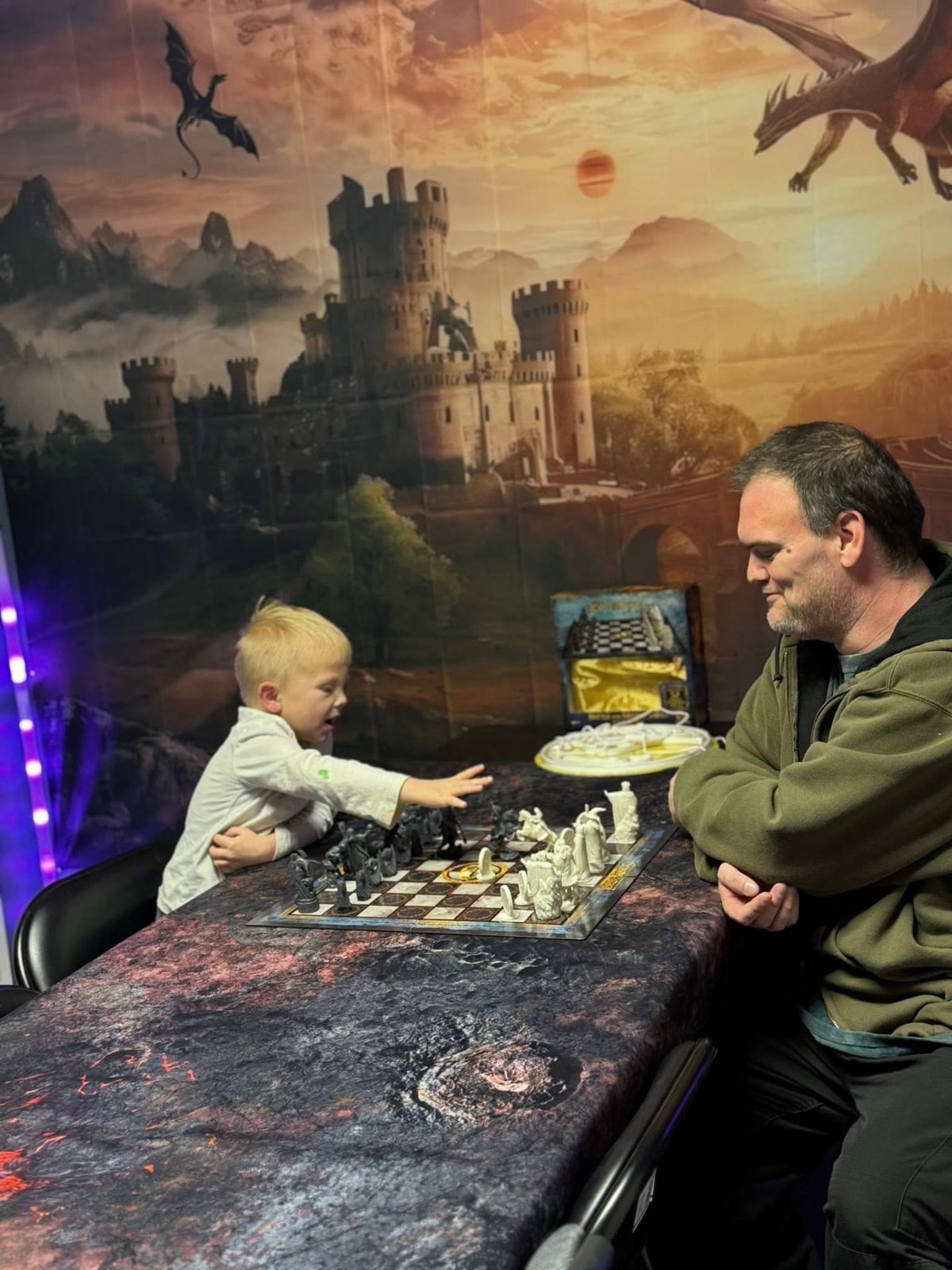 Young player reaching for a fantasy chess piece across the board from an adult at the castle mural table