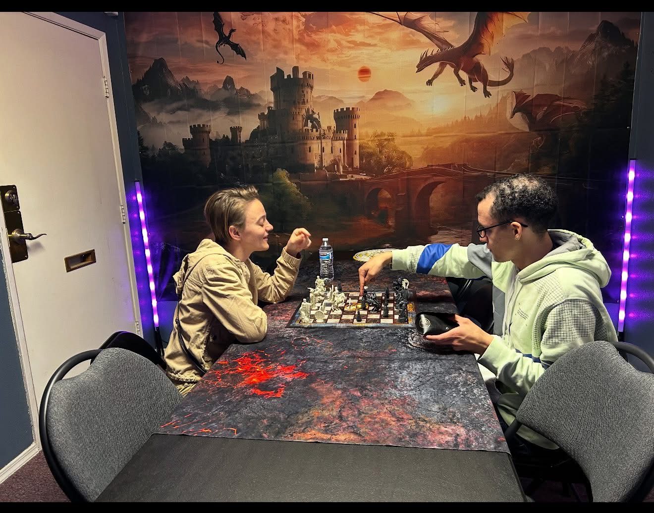 Two players facing off over a fantasy chess set at Nightfall Games™ in Pahrump, with the dragon and castle mural glowing behind them