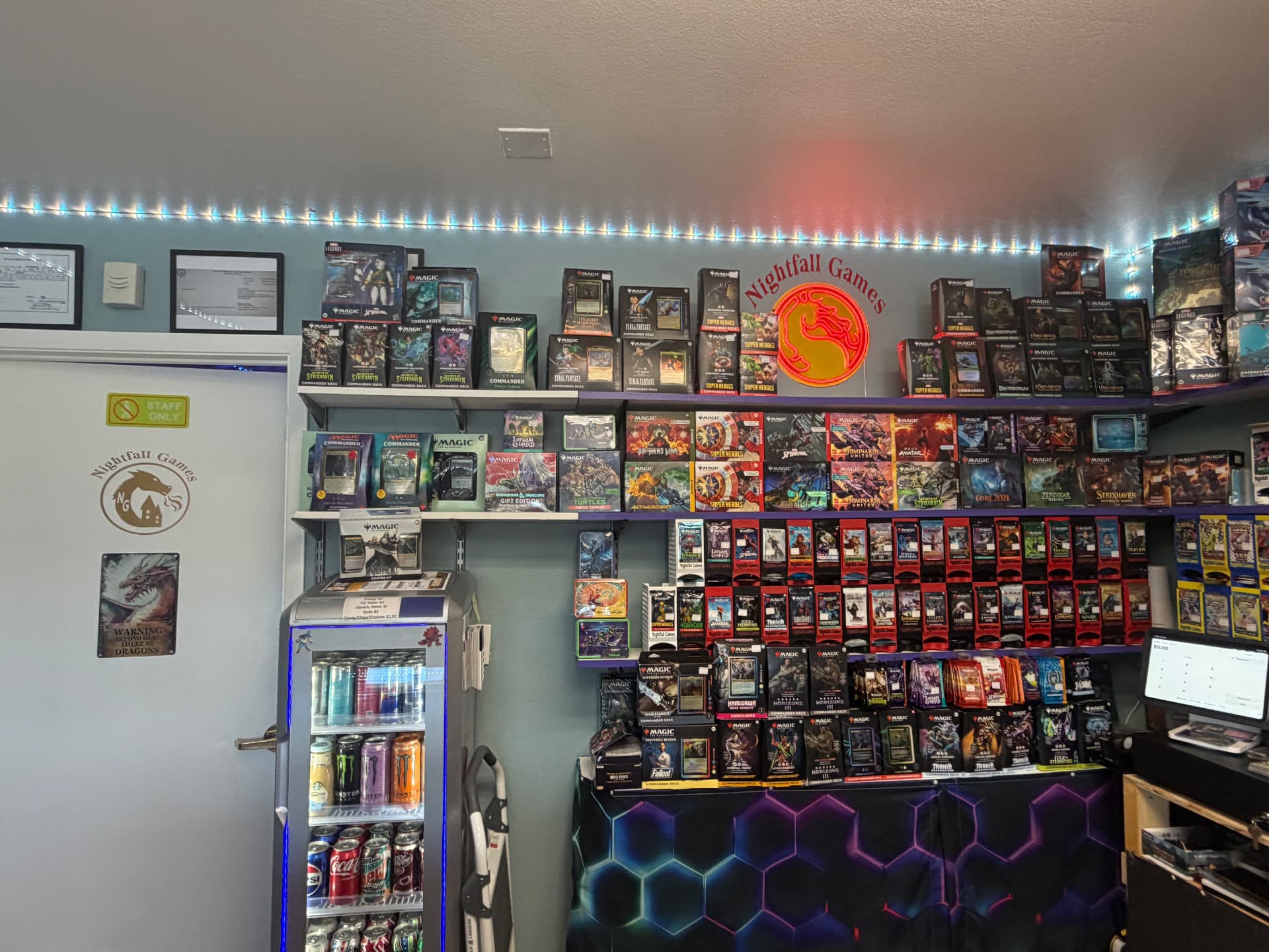 Magic: The Gathering wall at Nightfall Games™ lined with Commander decks, bundles and booster packs under the glowing Nightfall Games™ dragon sign