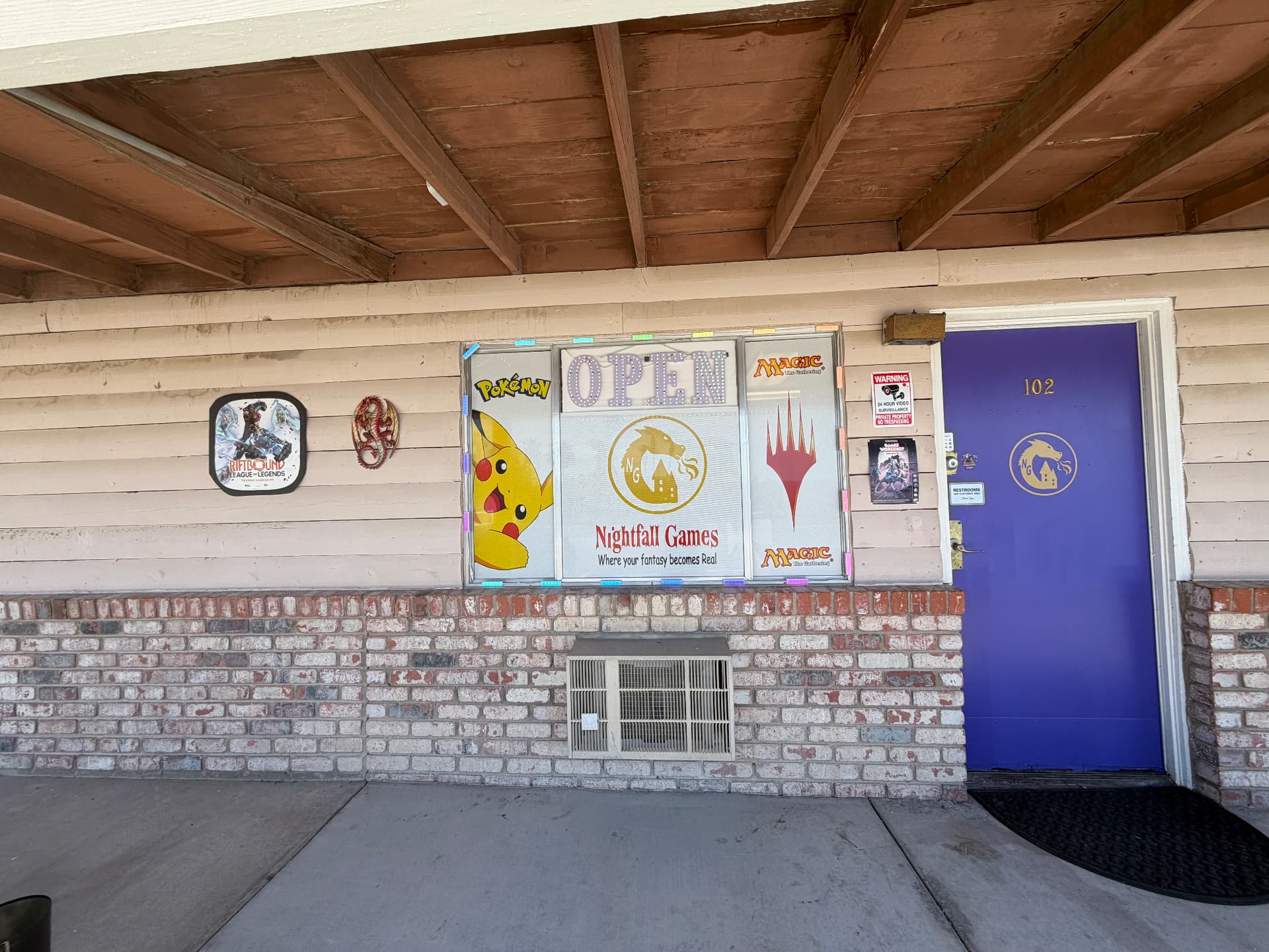 Entrance to Nightfall Games™ at 1201 S NV-160 Unit 102 in Pahrump, Nevada, with the OPEN sign, gold dragon window logo and Pokémon, Magic and Riftbound window art