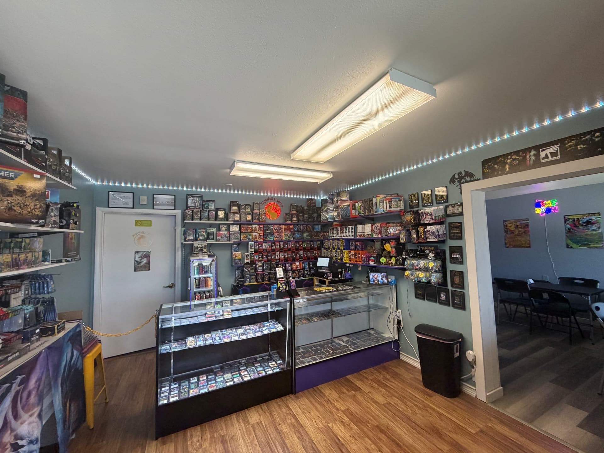 Wide view of the Nightfall Games™ sales floor with the checkout counter, trading card singles cases, the sealed product wall and the doorway into the game room