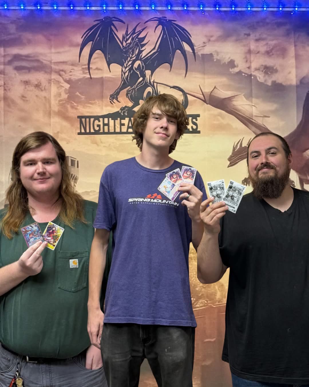 Three One Piece Card Game players holding their prize cards in front of the Nightfall Games™ dragon banner after a tournament in Pahrump