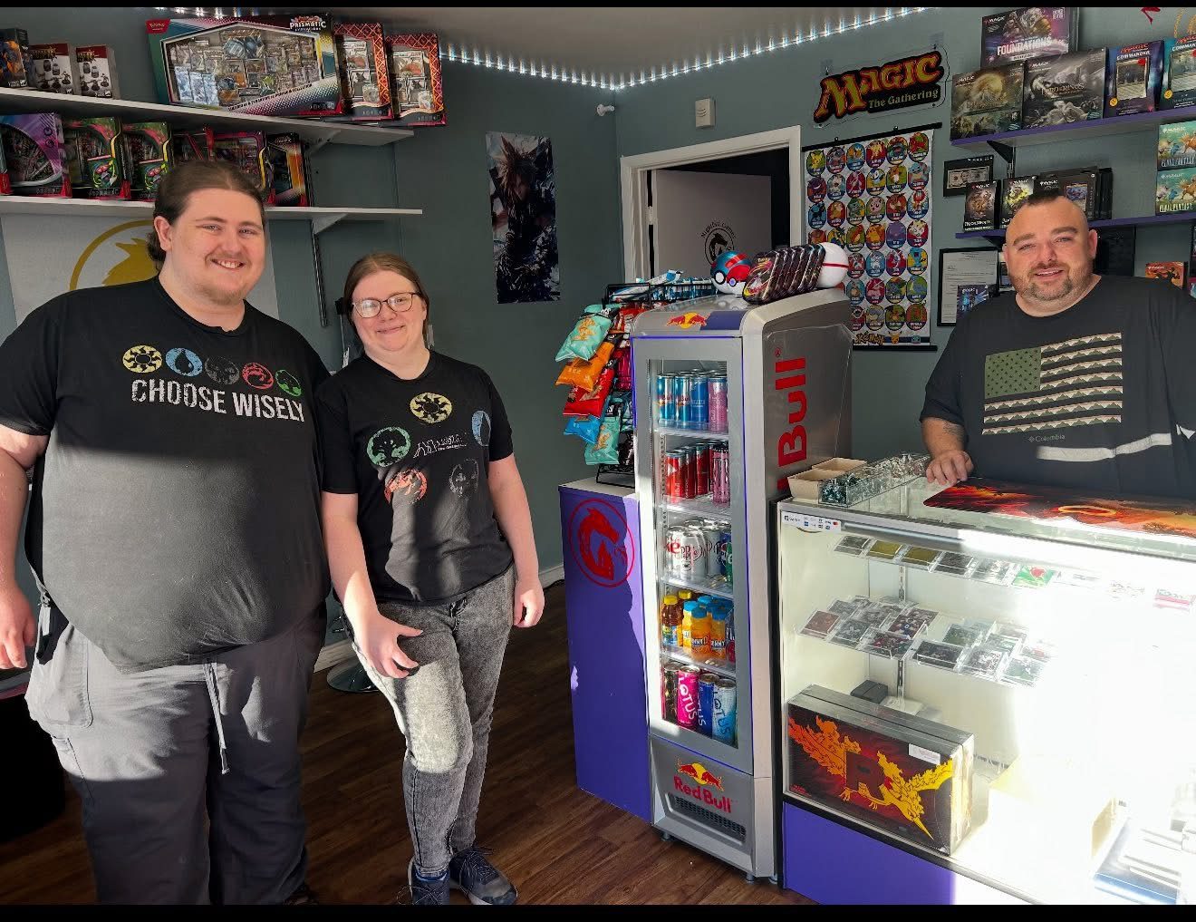 Three Nightfall Games™ team members smiling by the front counter, singles case and Magic display wall