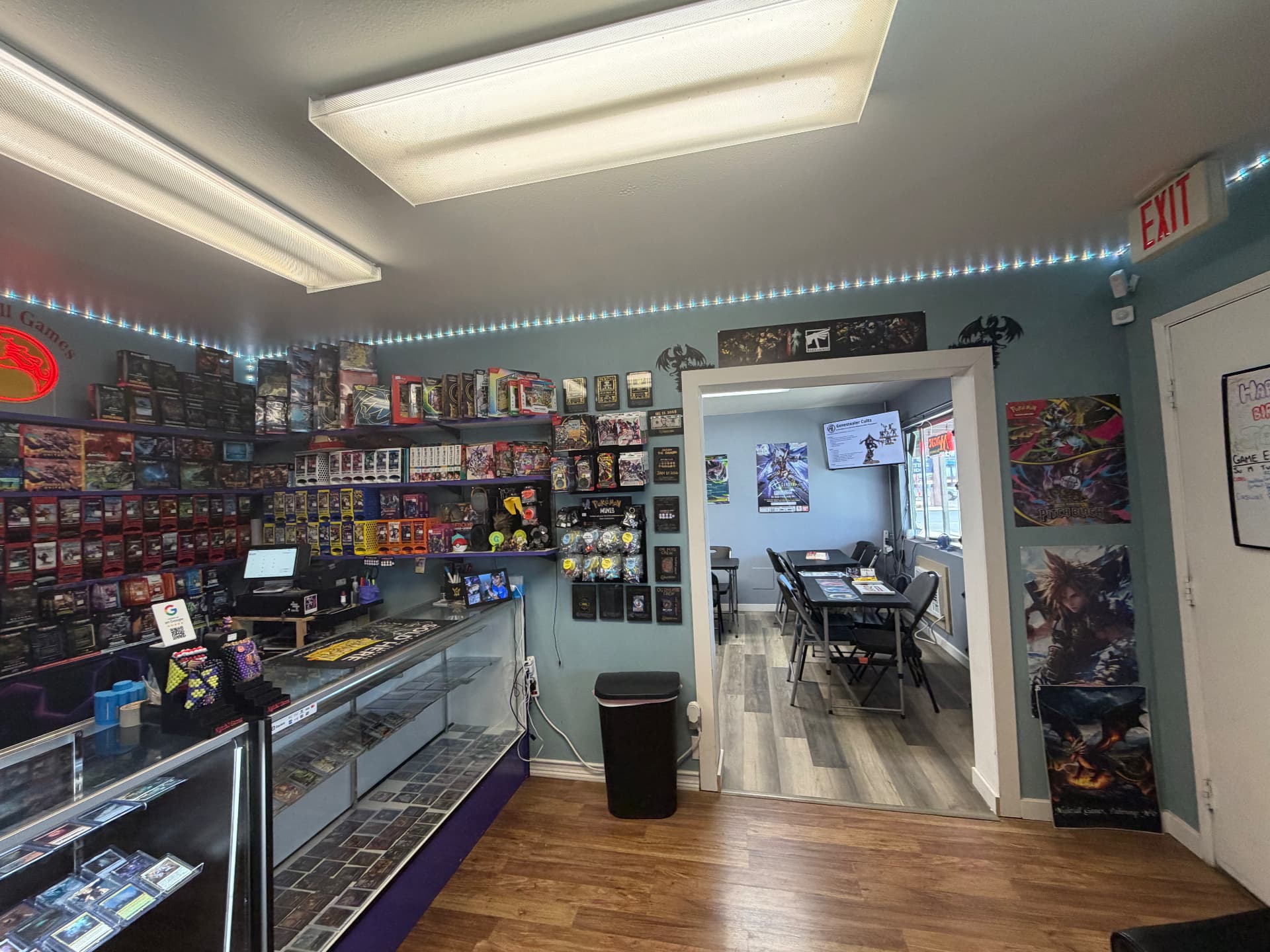Front counter at Nightfall Games™ with trading card singles in glass cases, sealed product on the shelves and the doorway through to the game room