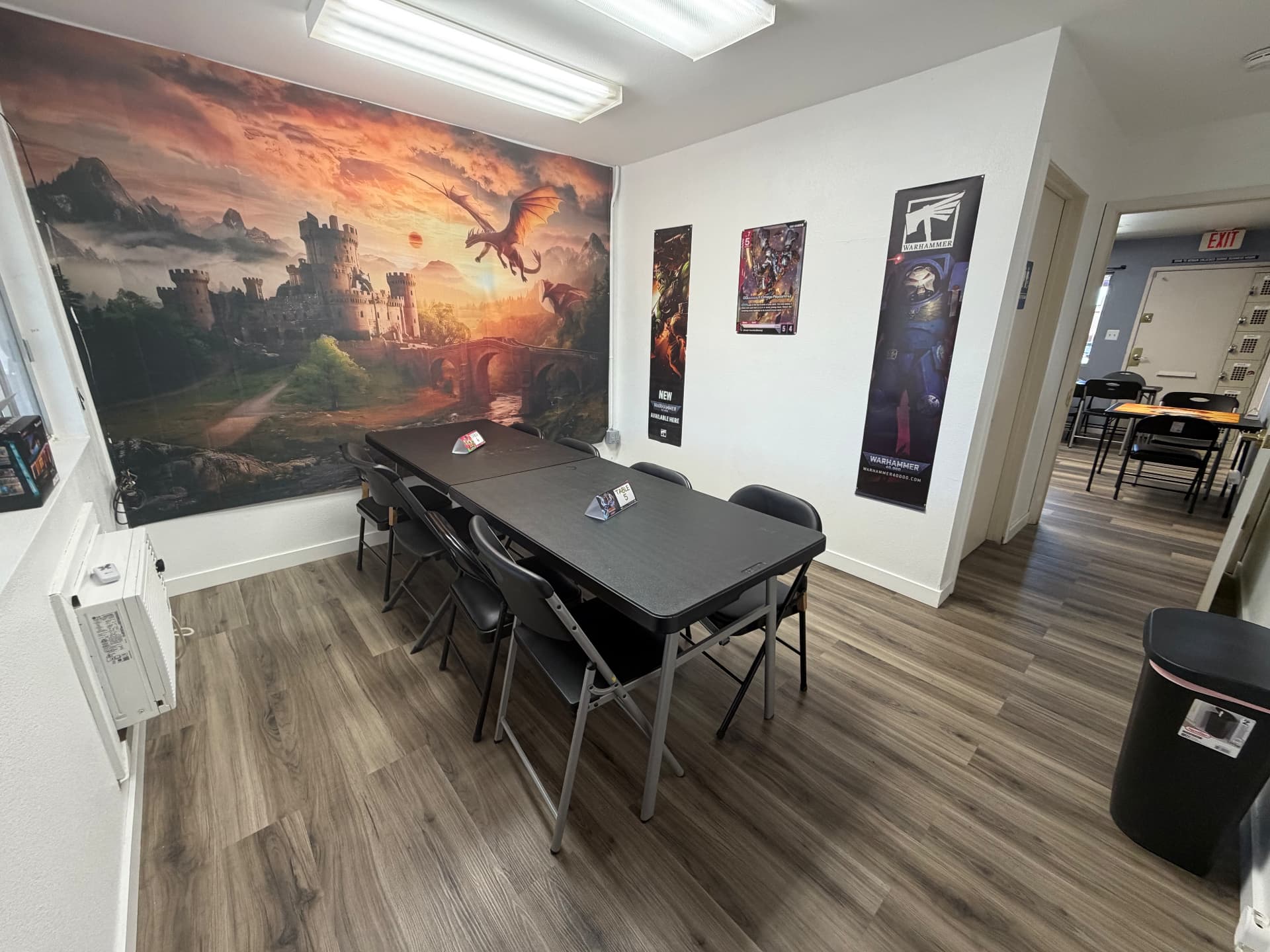 Castle and dragon mural wall in the Warhammer play room at Nightfall Games™, with a six-seat table and Warhammer 40,000 banners