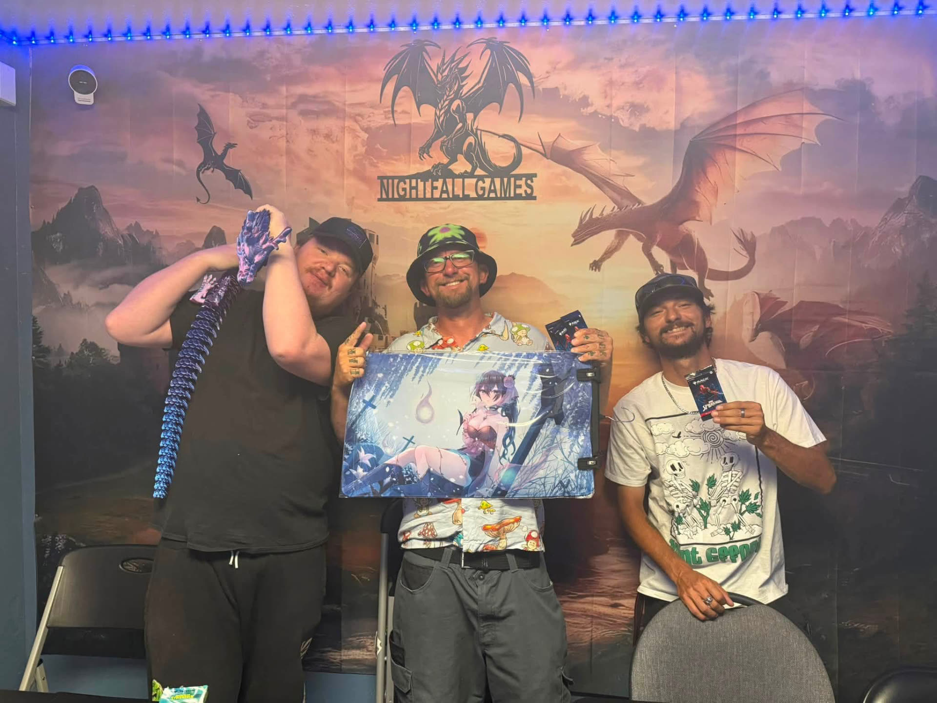 Three Nightfall Games™ players holding a playmat, booster packs and an articulated dragon in front of the store's dragon and castle mural