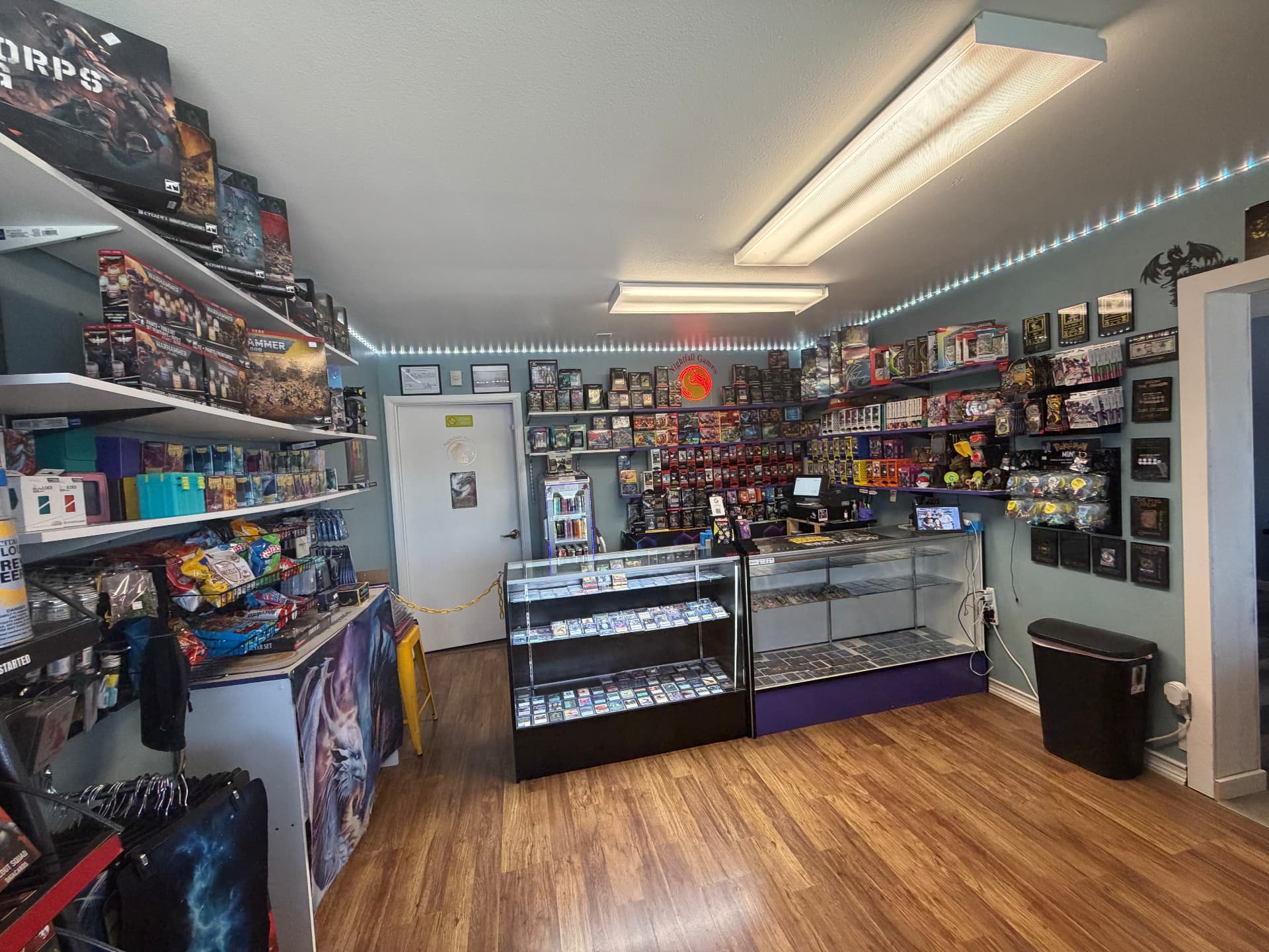 Nightfall Games™ sales floor seen from the Warhammer shelving, looking across the singles cases toward the sealed product wall