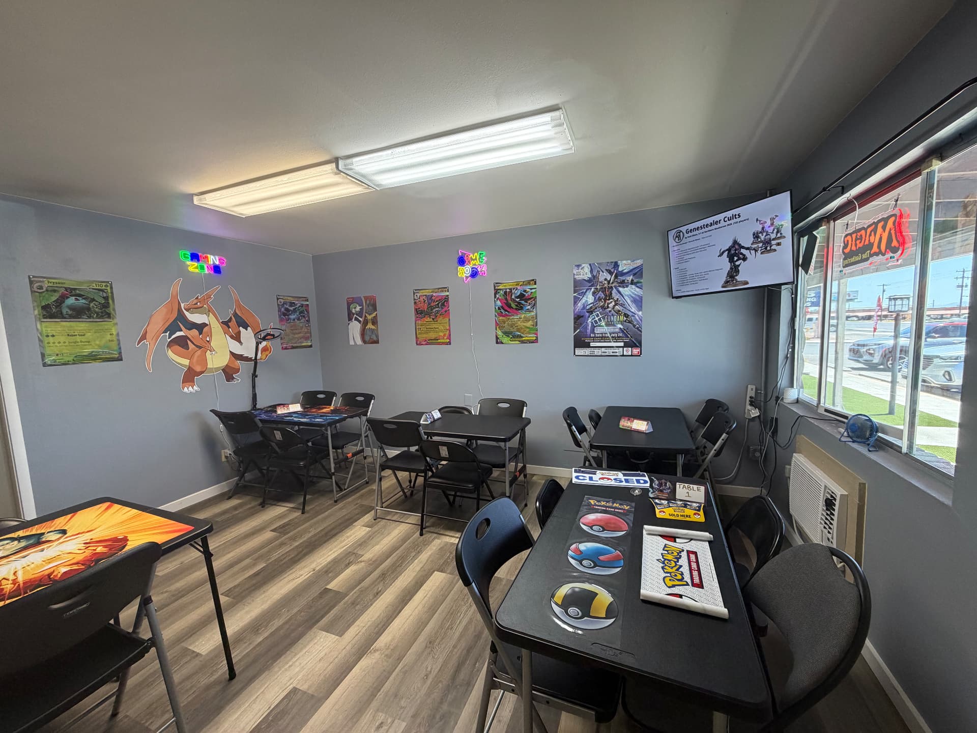Game room at Nightfall Games™ with Pokémon playmats on the tables, a Charizard cutout, Gaming Zone neon sign and a Gundam release poster