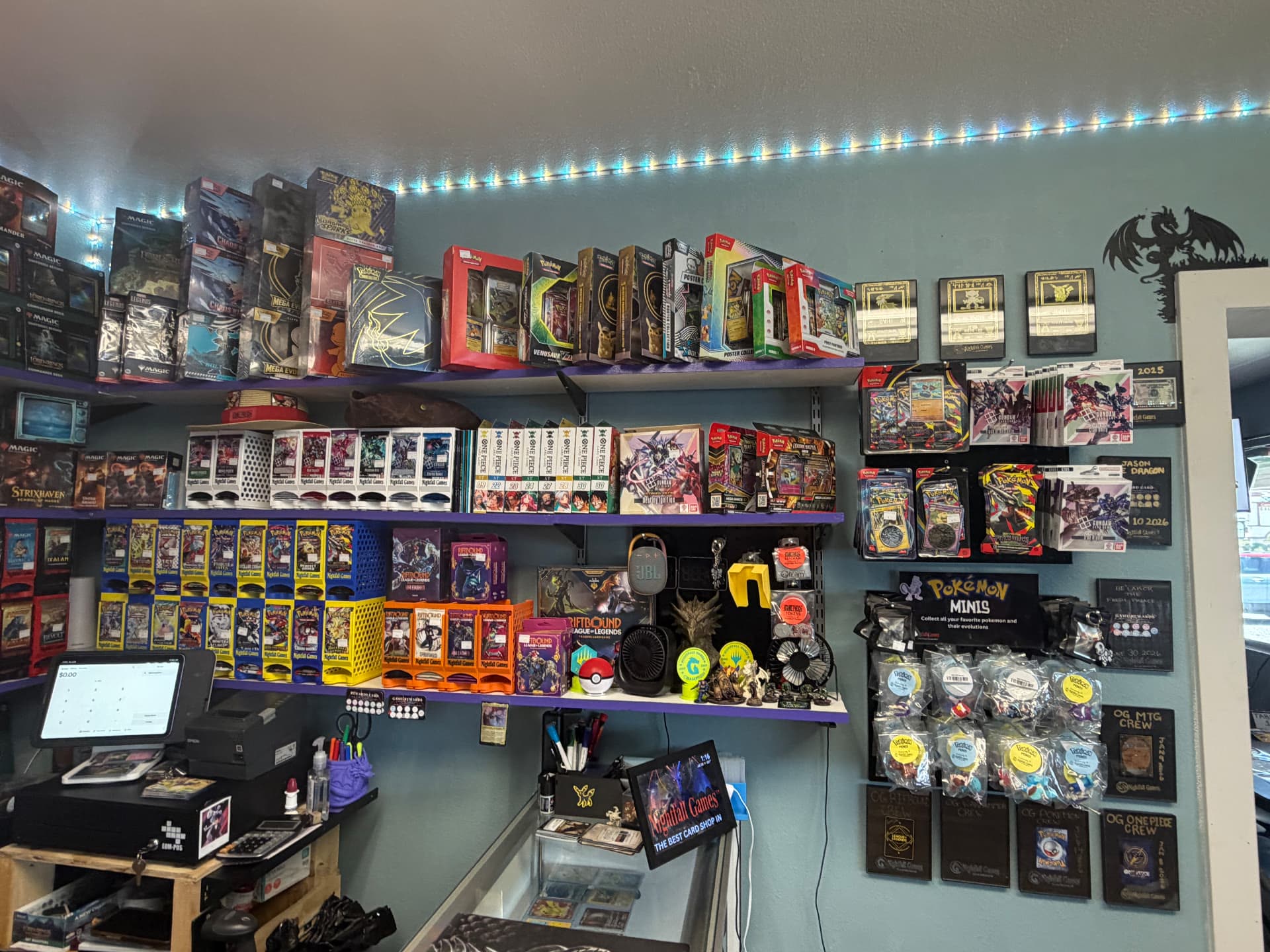 Purple shelving wall at Nightfall Games™ stocked with Pokémon, One Piece, Gundam, Riftbound and Magic sealed product beside the Pokémon Minis display