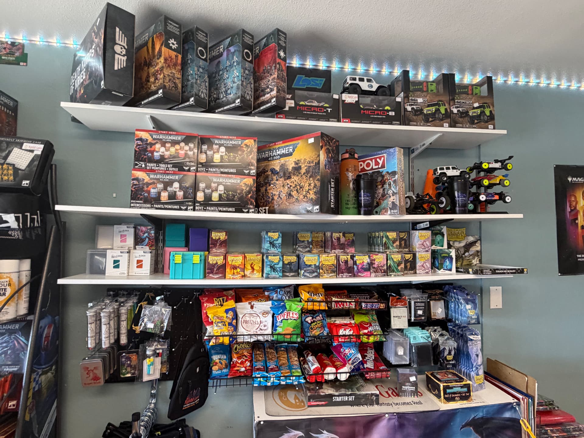 Shelves at Nightfall Games™ holding Warhammer 40,000 starter and paint sets, RC crawler kits, Dragon Shield sleeves, deck boxes and snacks