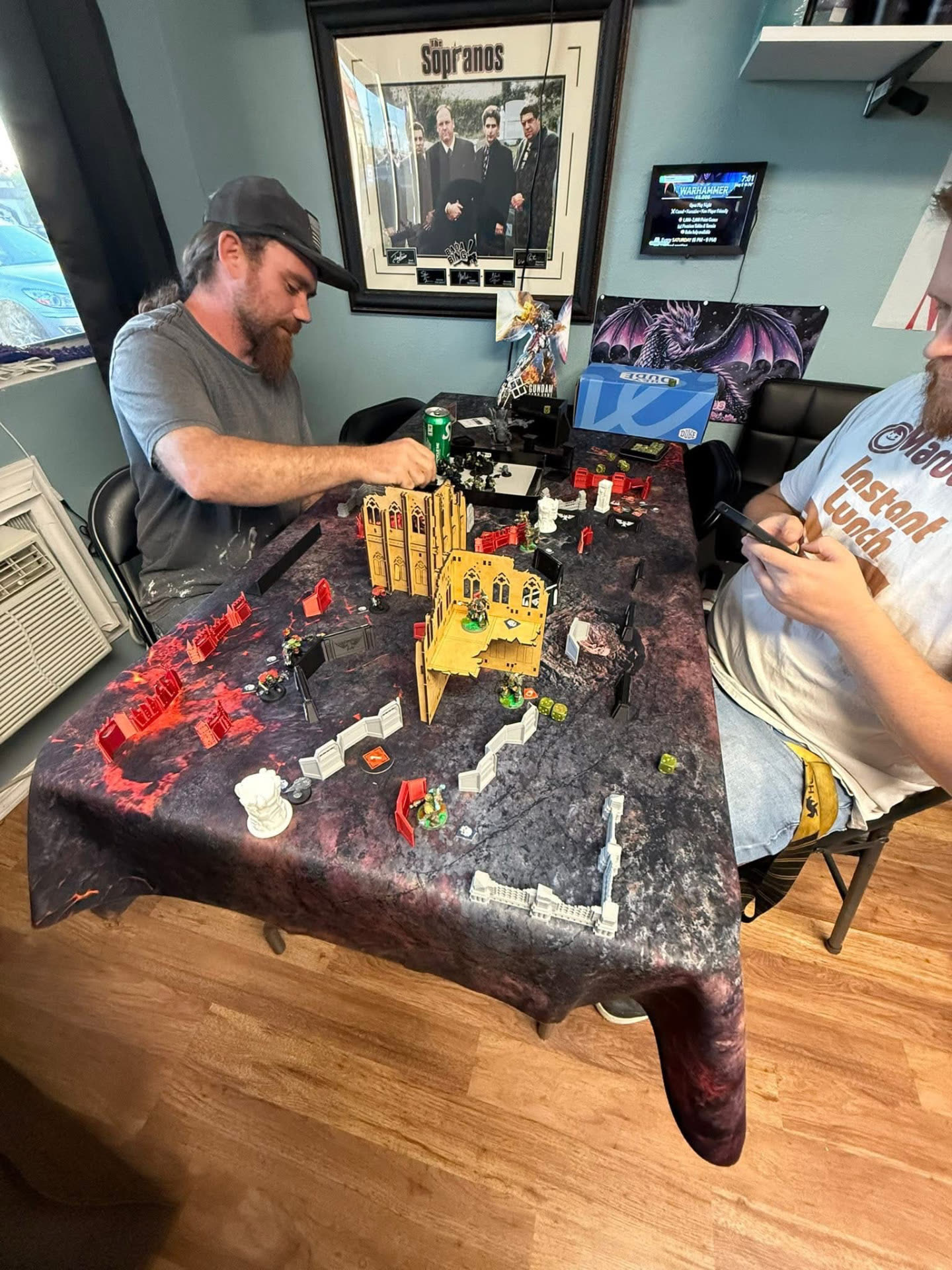 Two players mid-game on a Warhammer 40,000 battle mat with painted miniatures and ruined terrain at Nightfall Games™